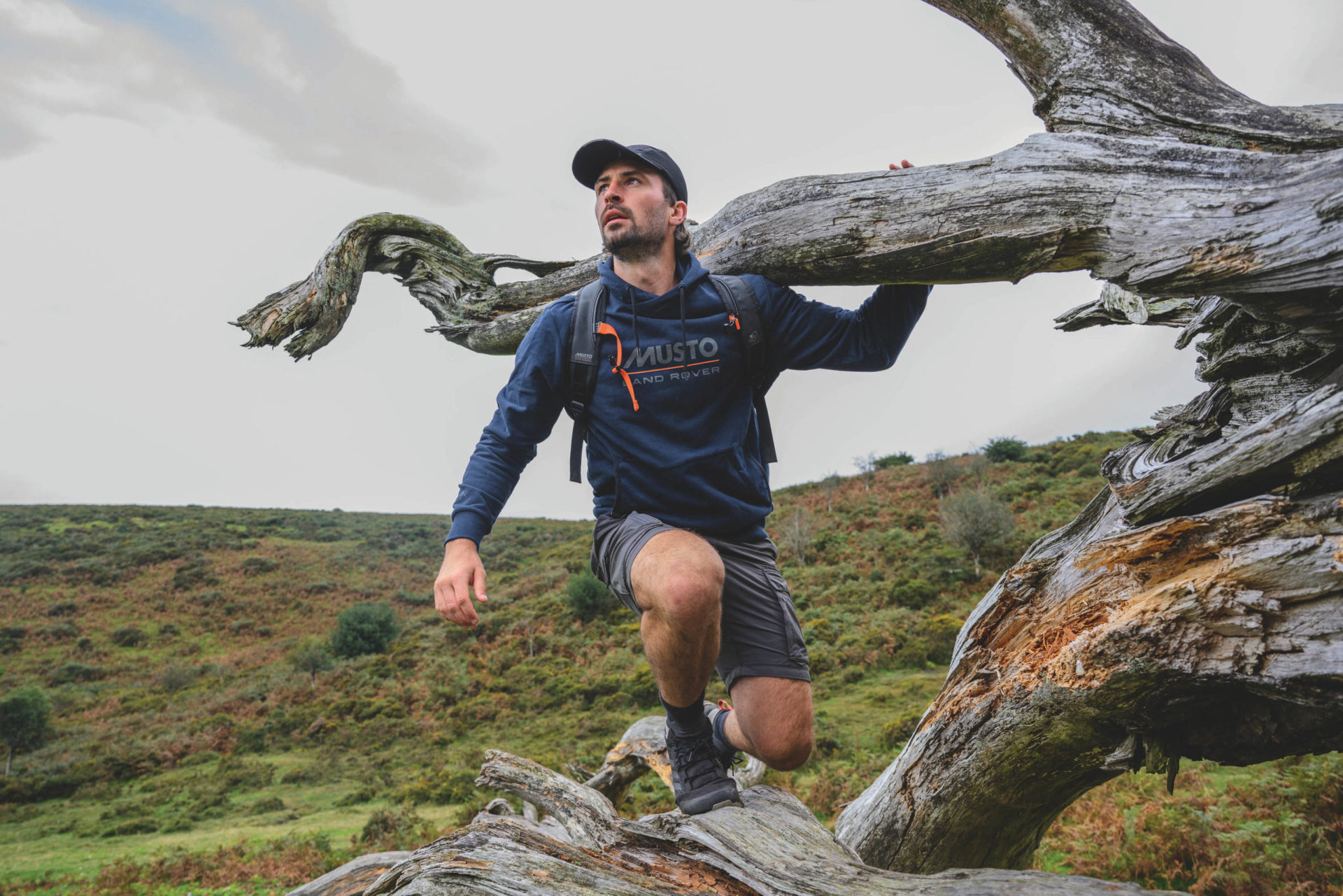 Man in Musto jacket and shorts on tree. Background: fields and cloudy sky.