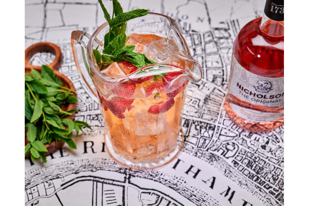 A jug of orange liquid next to a basket of herbs and a bottle of Nicholson Blood Ornage Gin