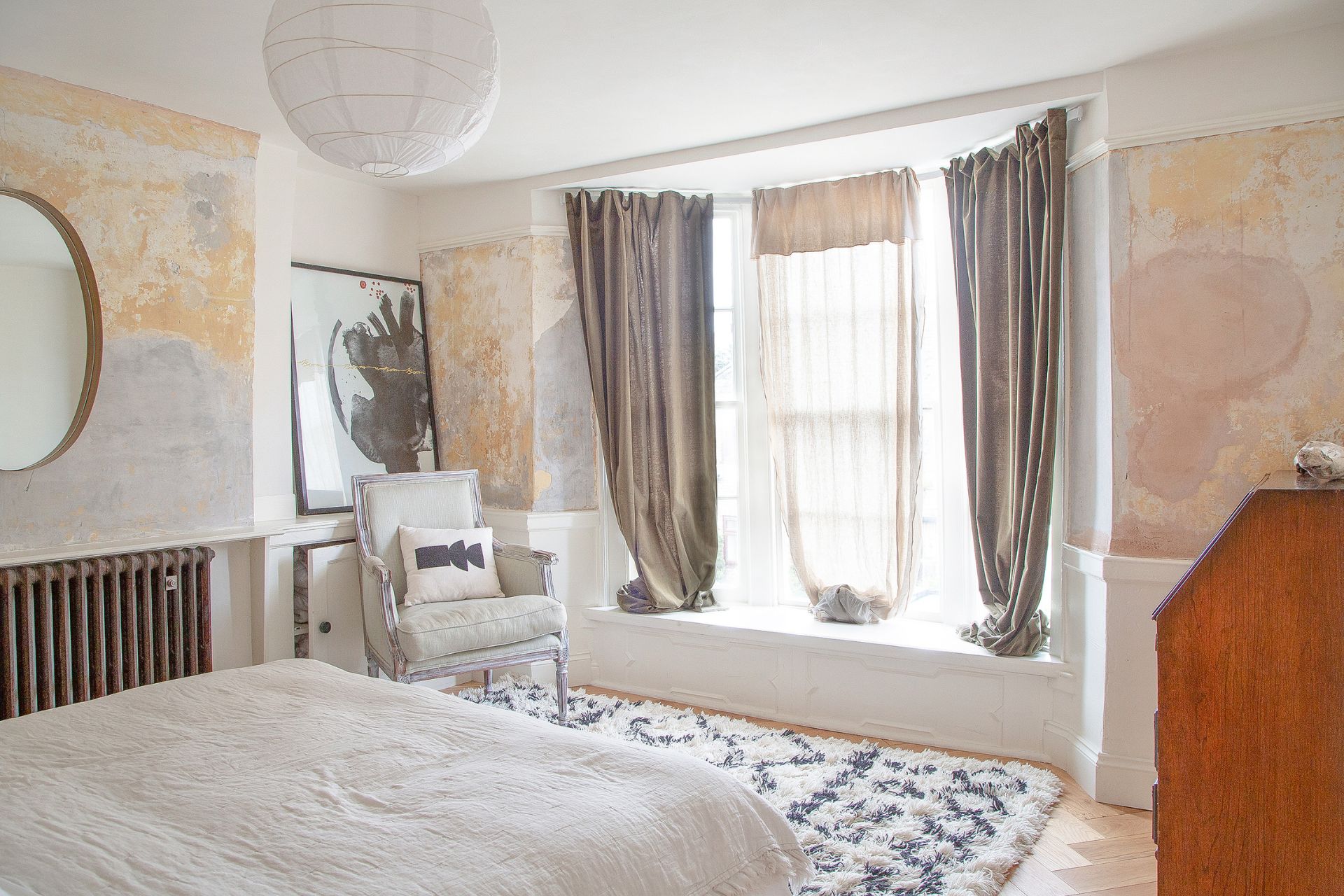 Modern bedroom with a bay window, taupe curtains, a round gold mirror and a wooden dresser.