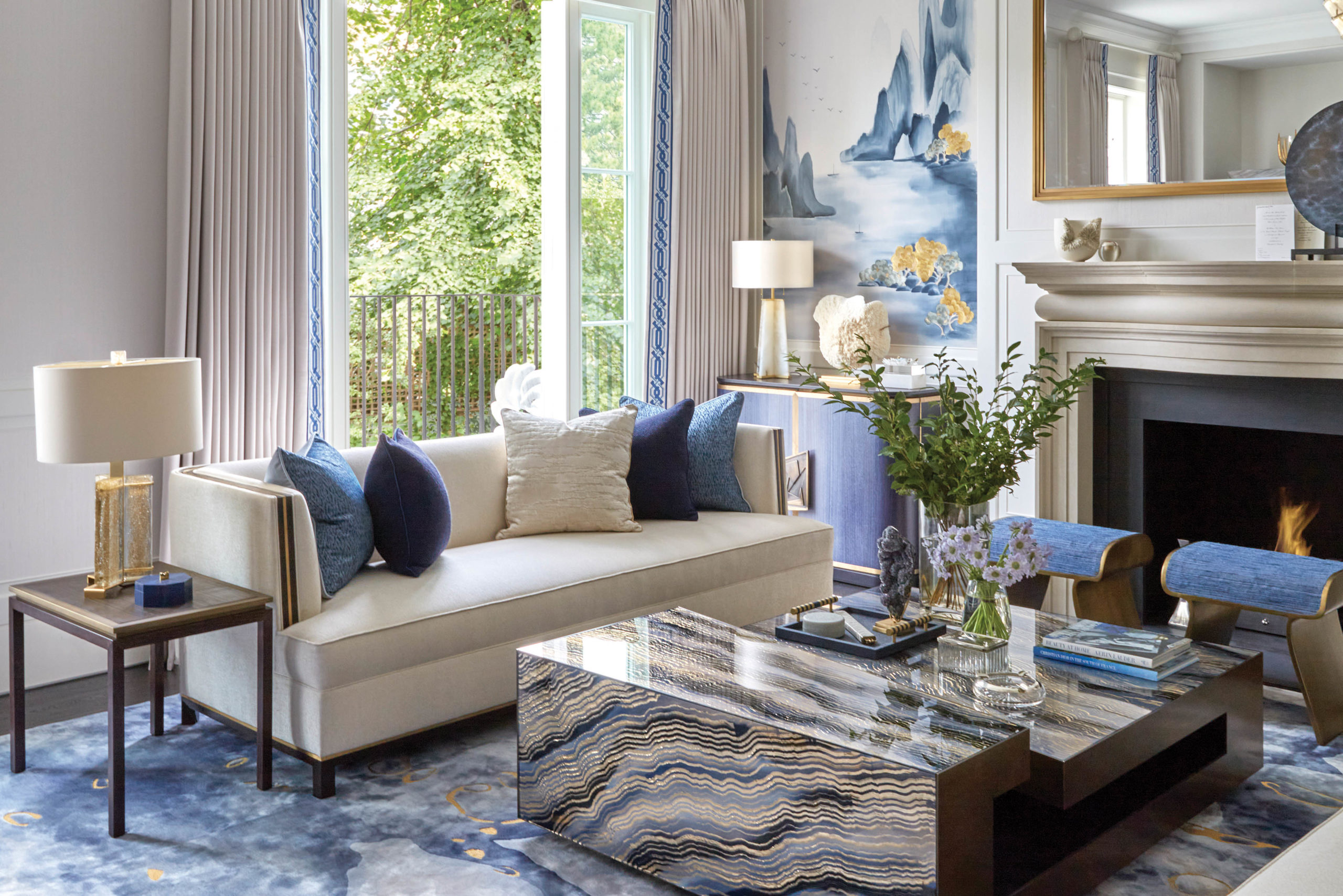 Katharine Pooley designed blue and white sitting room. Foreground: blue swirl carpet, metallic coffee table, white sofa with blue scatter cushions. Background: white fireplace, windows.