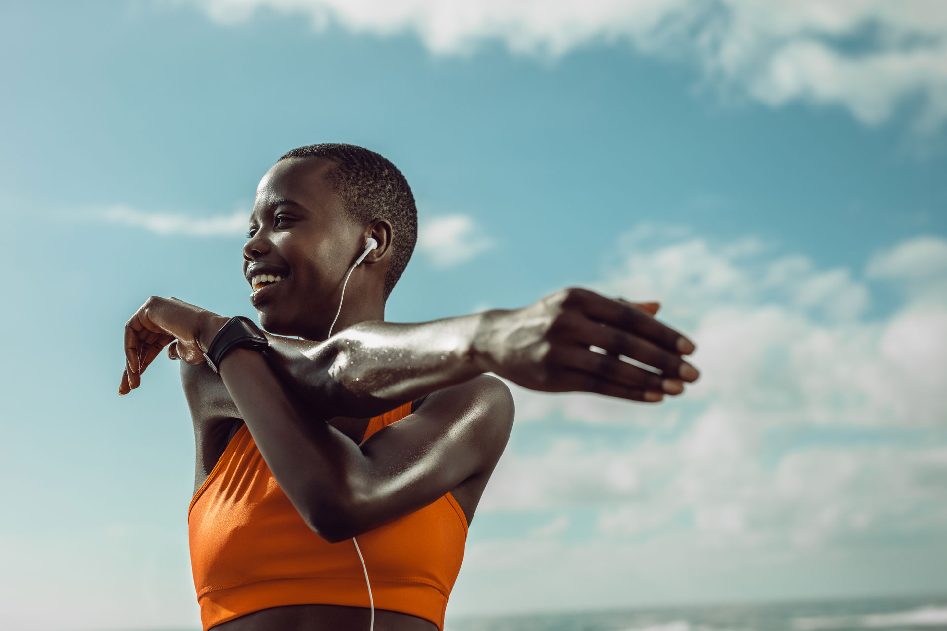 a black woman with an orange sports bra stretches her arms in a blue sky backdrop