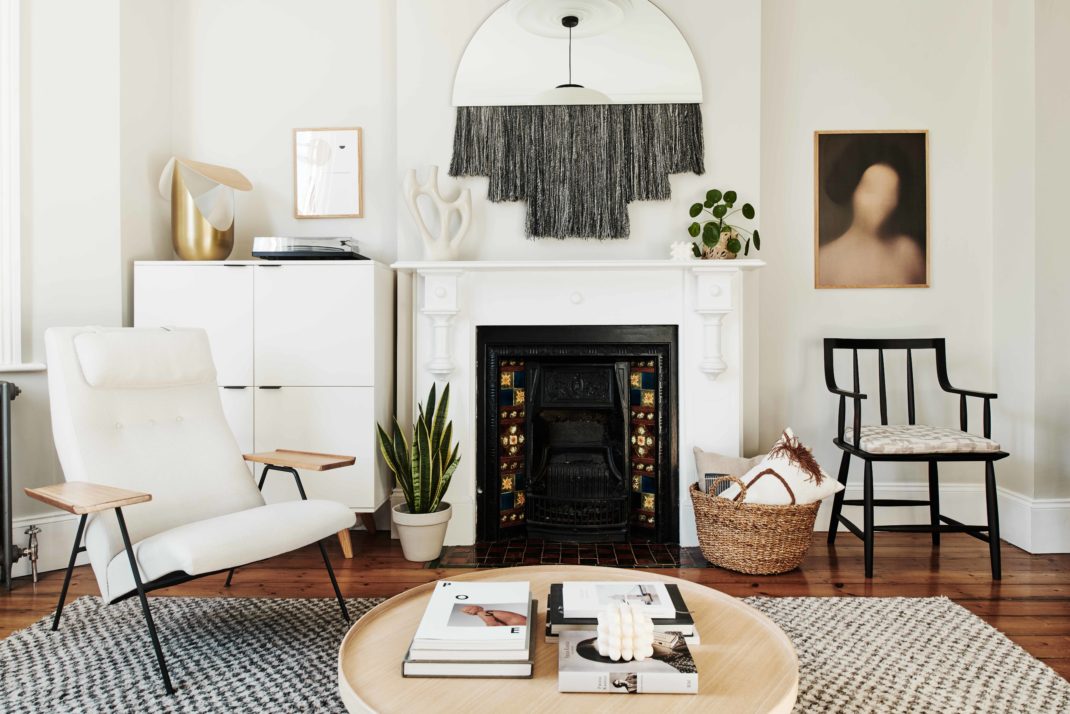 White sitting room with monochrome rug, wooden coffee table and white modernist chair. In the background: white fireplace, mirror with fringed detailing, modern ornaments.