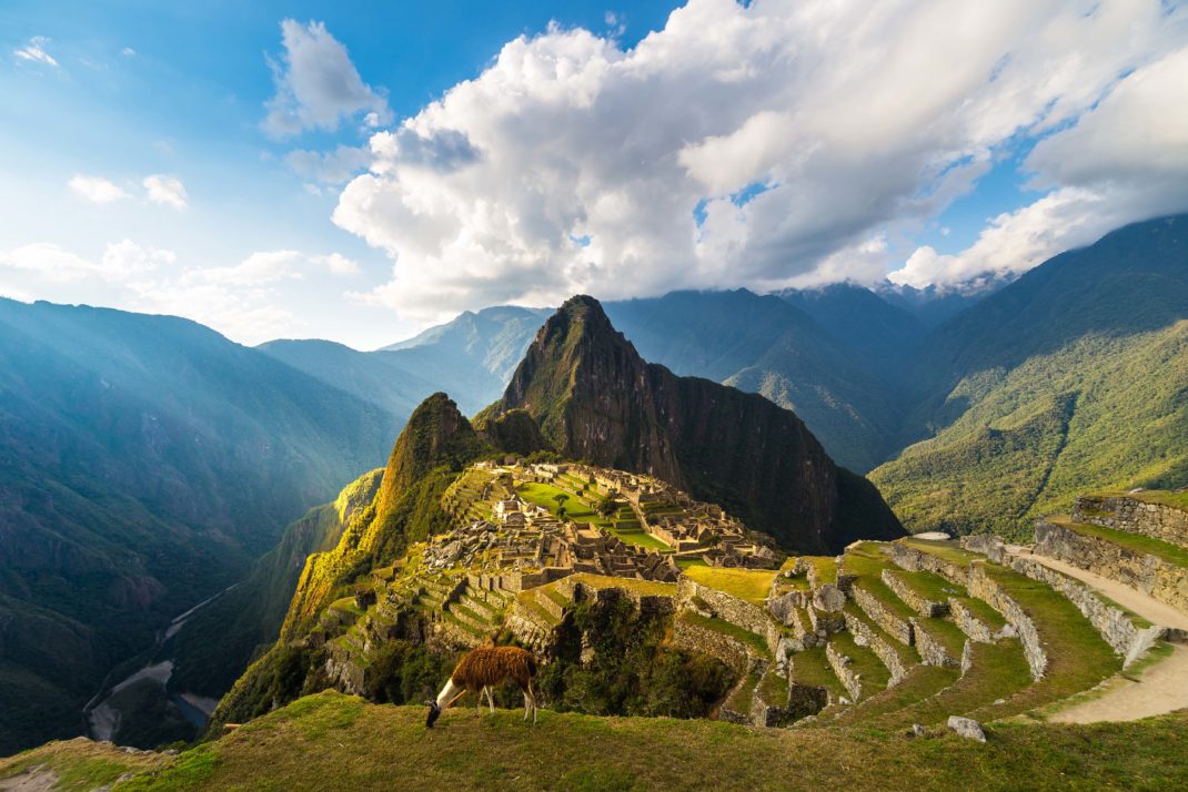 Machu Picchu illuminated by the warm sunset light. Wide angle view from the terraces above with scenic sky and sun burst. Dreamlike travel destination, world wonder. Cusco Region, Peru.