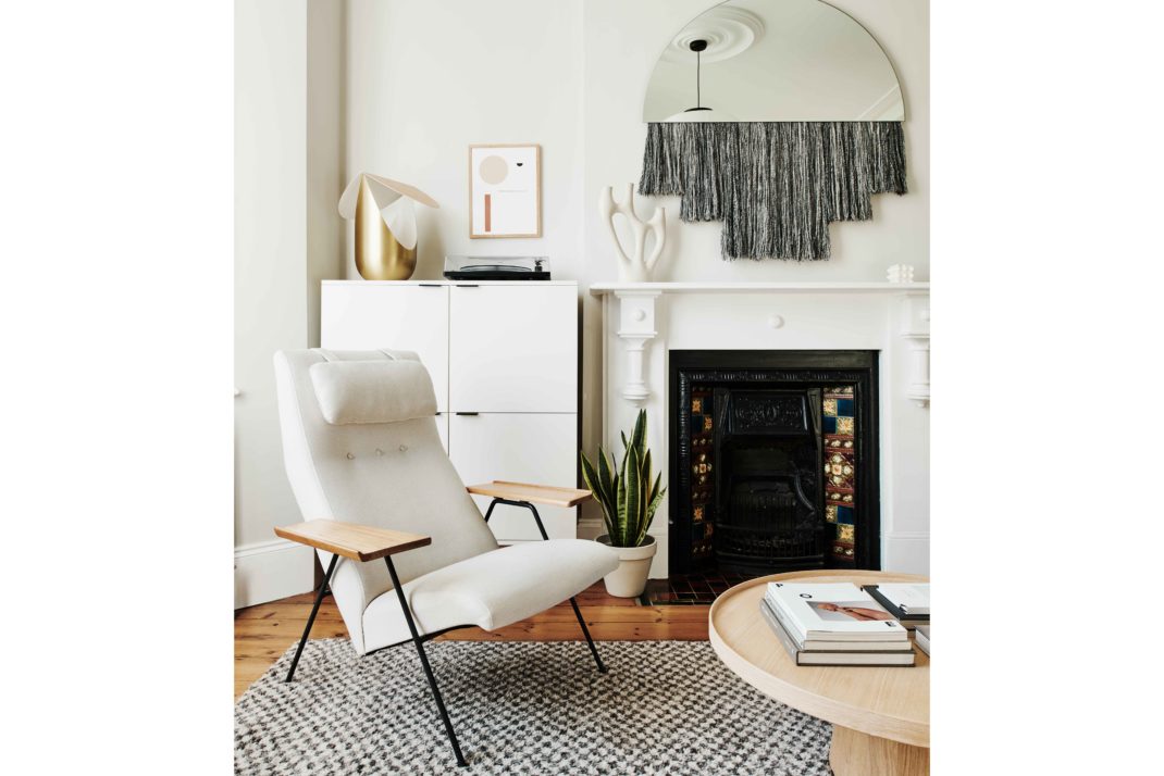 White sitting room with monochrome rug, wooden coffee table and white modernist chair. In the background: white fireplace, mirror with fringed detailing, modern ornaments.