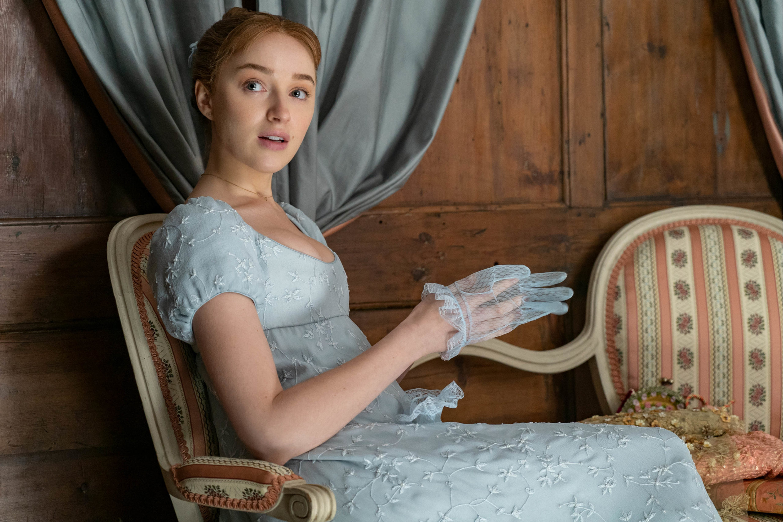 Daphne Bridgerton from Netflix Bridgerton, seated in floral patterned chair. Background: wood panelling, blue curtains