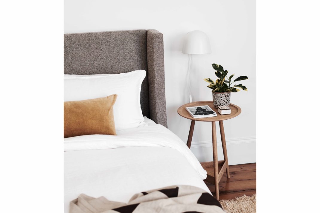 Modern bed with fawn bedstead, white bedding sheets and amber throw cushion. Wooden bedside table with plant.