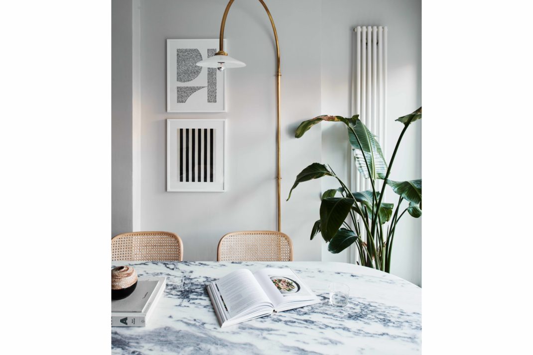 White painted kitchen with marble table. In the background: wicker chairs, brass lamp, plant, monochrome art prints.