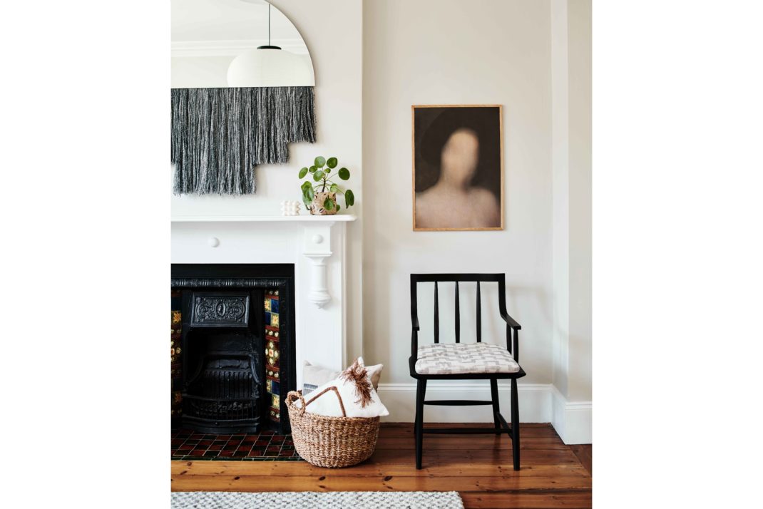 White modern sitting room with black chair, modern art print, mirror with grey fringe detail, white fireplace and basket.