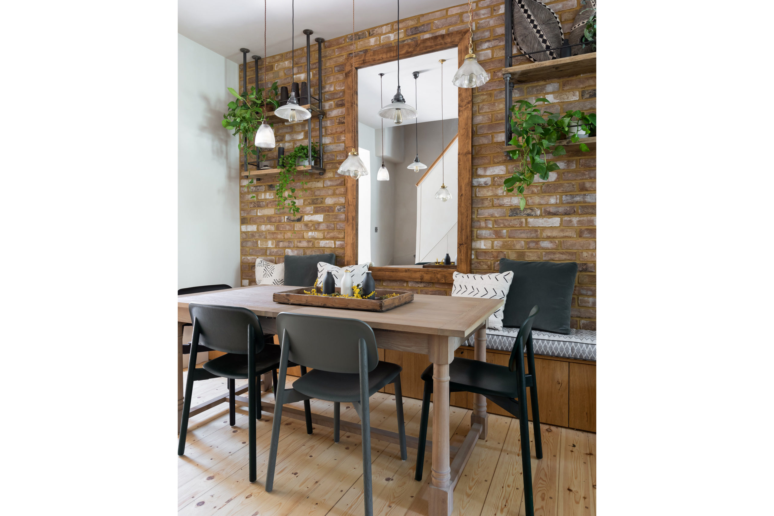 Dining area with exposed brick wall. Hanging pendant lights, four grey chairs around wooden table, hanging plants in background