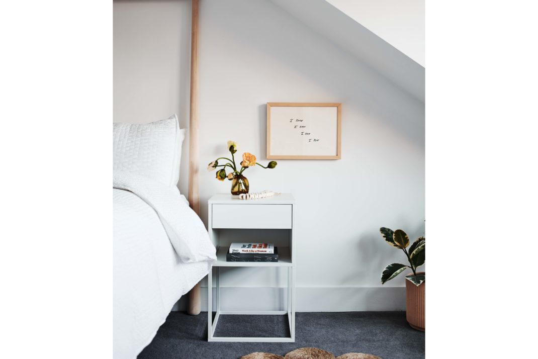 Bedroom with white bed and bedside table, grey carpet. In the background: plant, framed print.