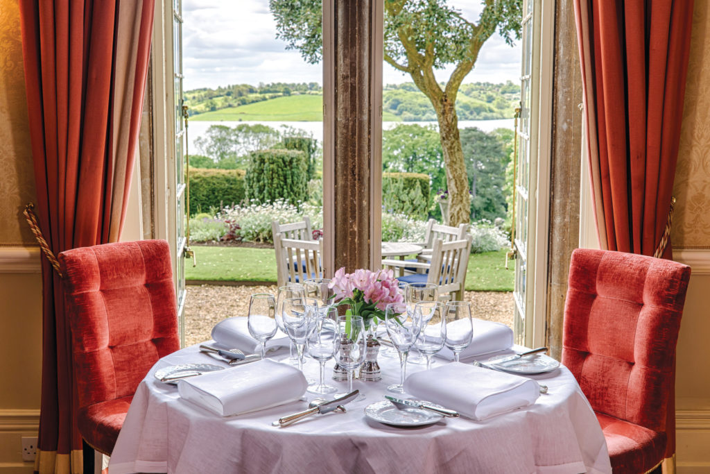 Table laid with white table cloth, red chairs and red curtains, window lading to garden