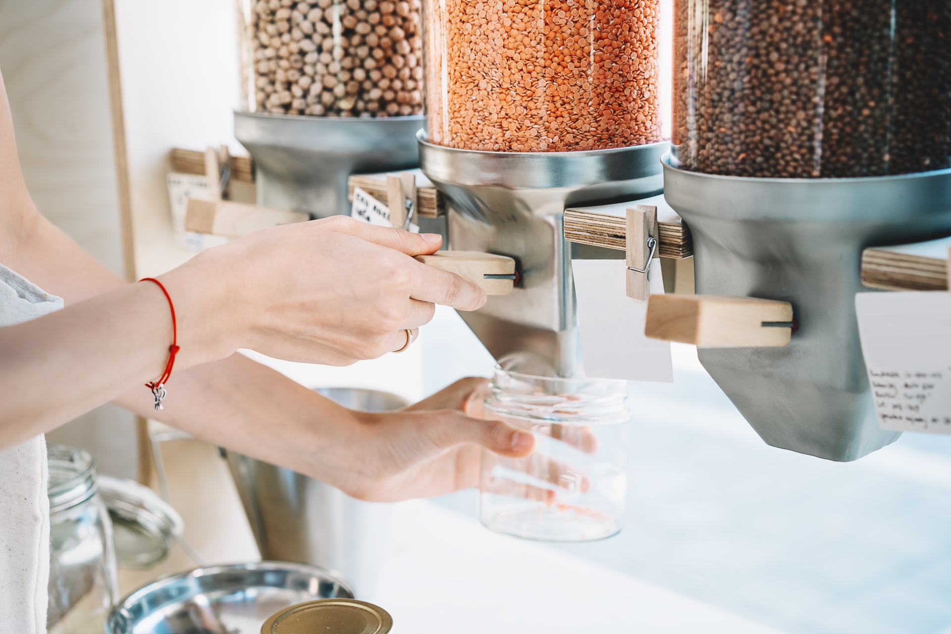 Refill Stores: A Guide - Sustainable shopping at small local businesses. Close-up image of woman pours red lentils in glass jar from dispensers in plastic free grocery store. Girl with cotton bag buying in zero waste shop. refilling jar with lentils