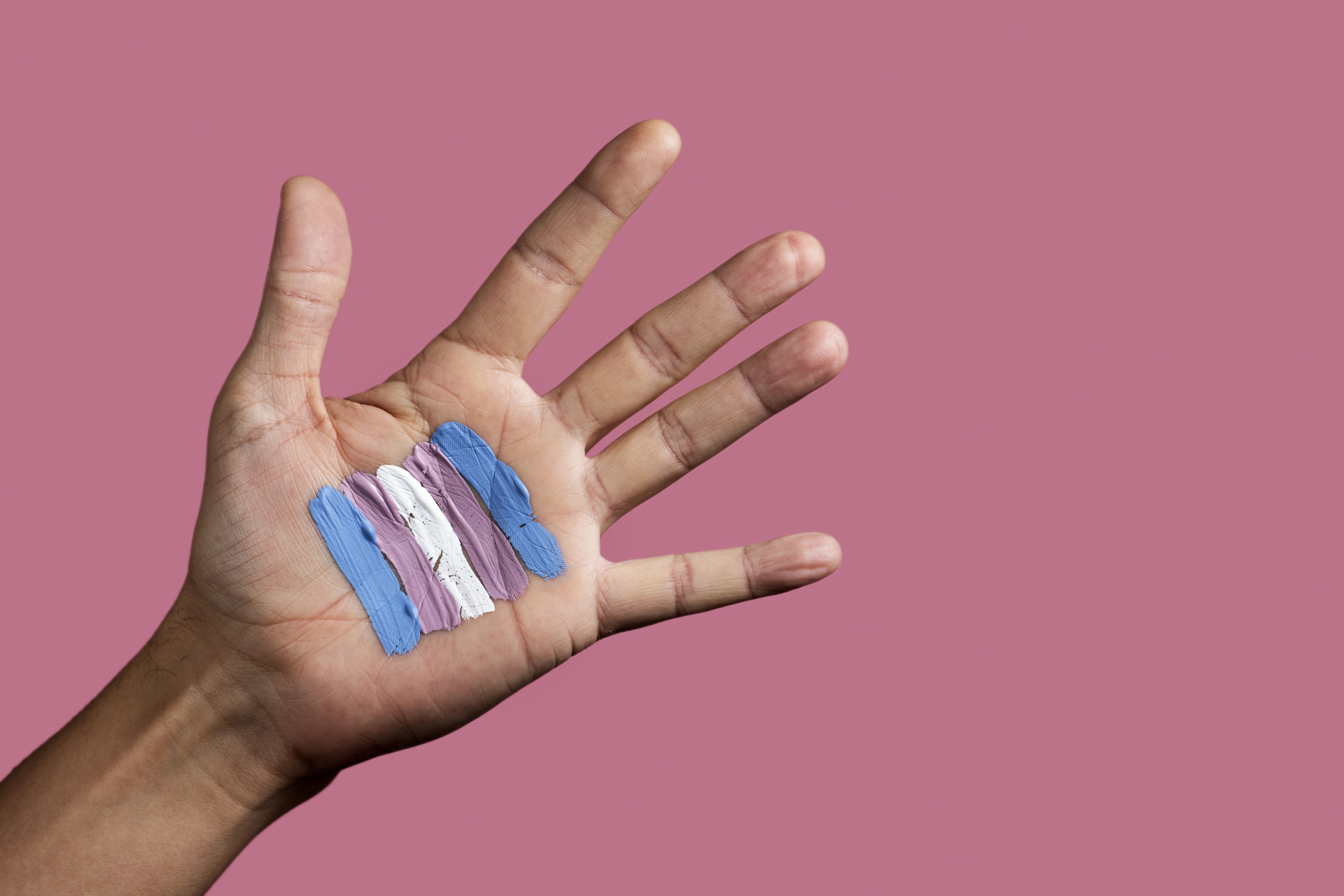closeup of the palm of the hand of a young caucasian person with a transgender flag painted in it, against a pink background with some blank space