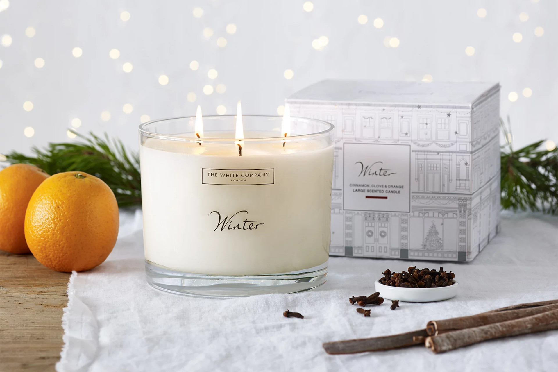 Candle on marble countertop next to oranges, cinnamon and a white box