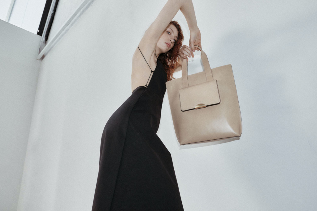 Woman in black gown holding cream handbag, stood in front of white wall