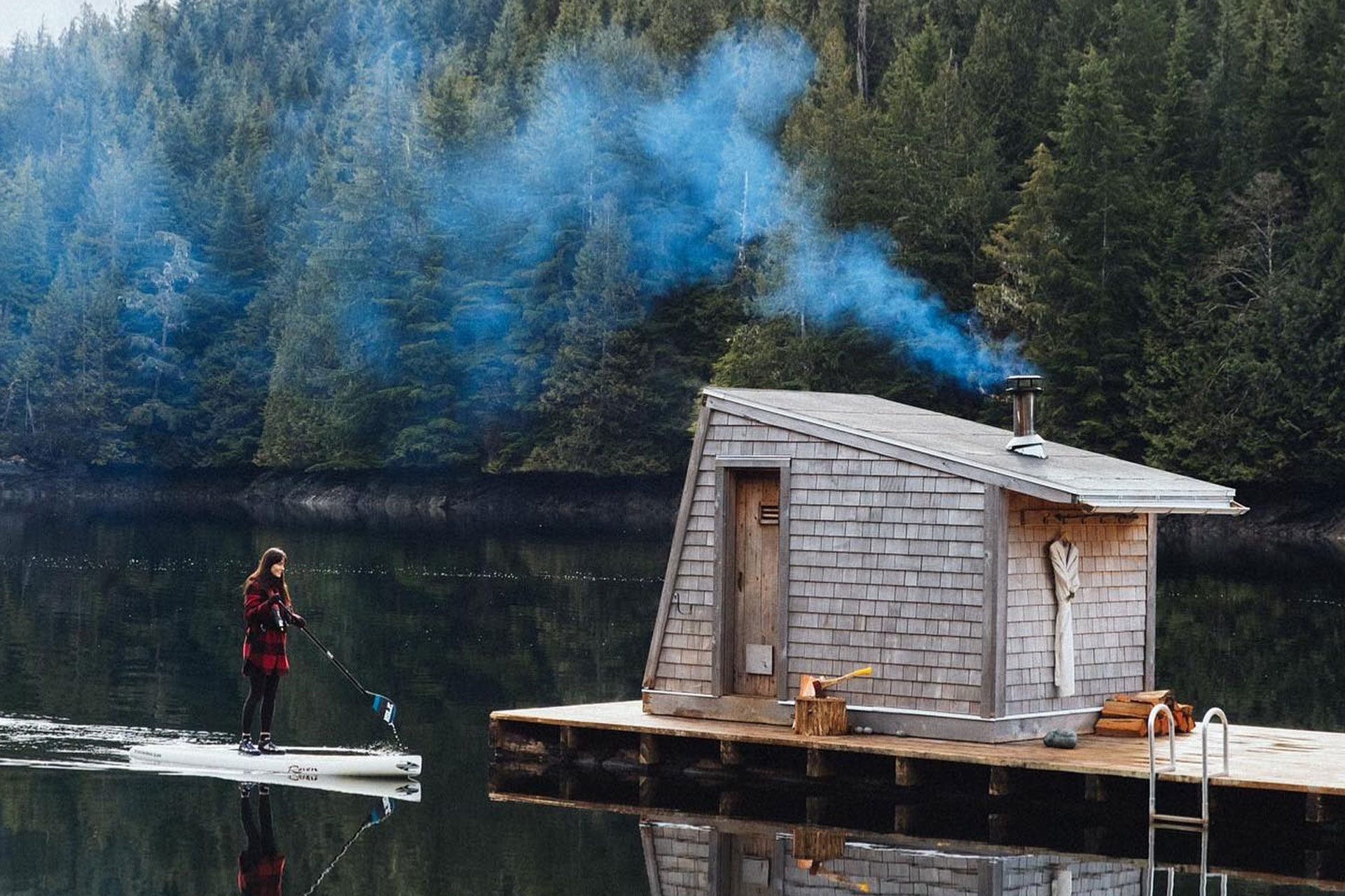 A woman oars to a floating sauna in a lake with trees