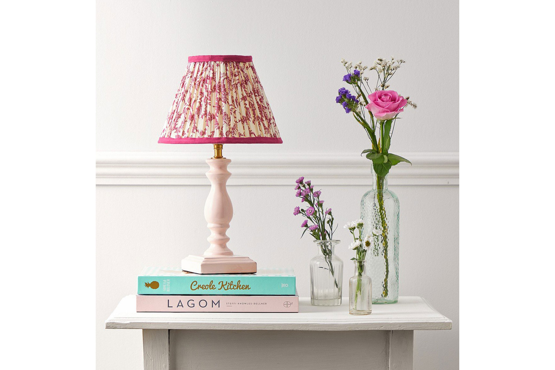 A chintzy pink linen Pooky lamp stands next to a pile of books and a vase of flowers