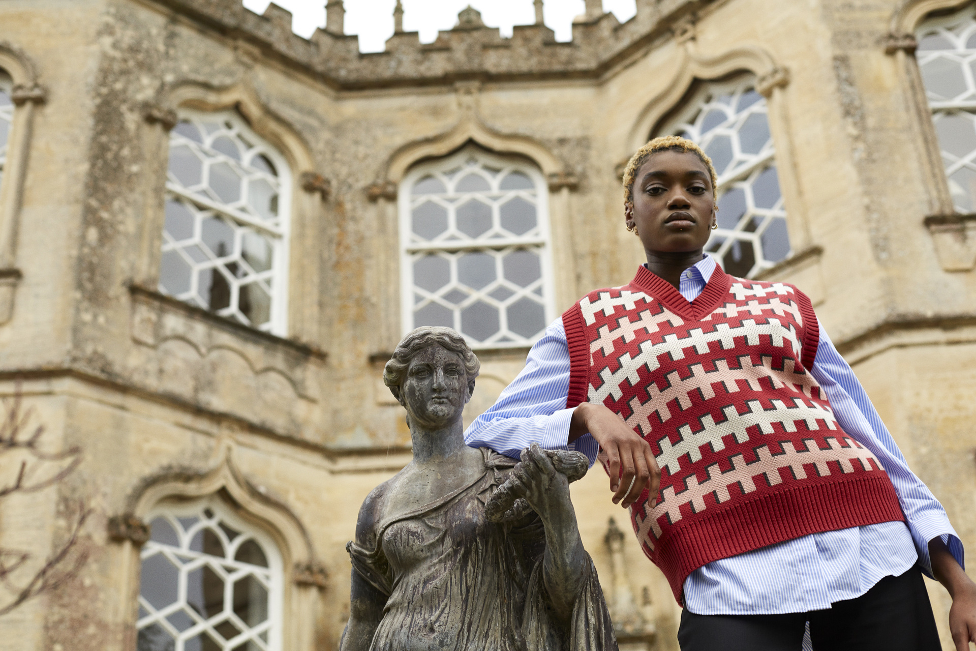 Model in red sweater vest standing next to a sculpture