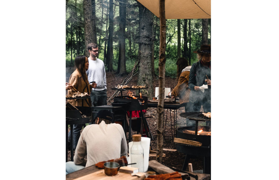 a group of people stand around a forest with an open fire