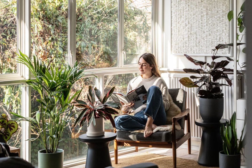 a girl sits amongst lots of potted plants beside a floor to ceiling window