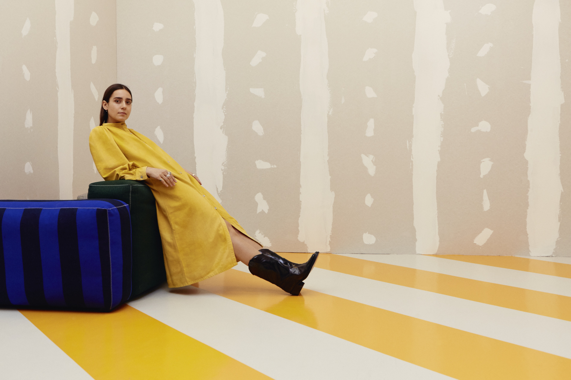 Woman in yellow dress reclining on blue stool, over yellow striped floor and beige striped wall