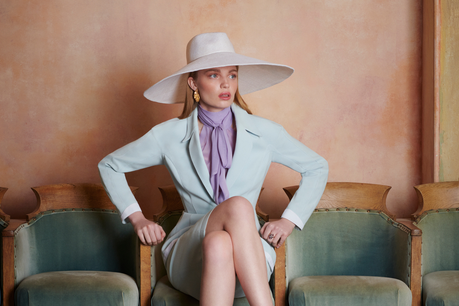 Woman in blue skirt suit and wide-brimmed hat sat on light green velvet chair row