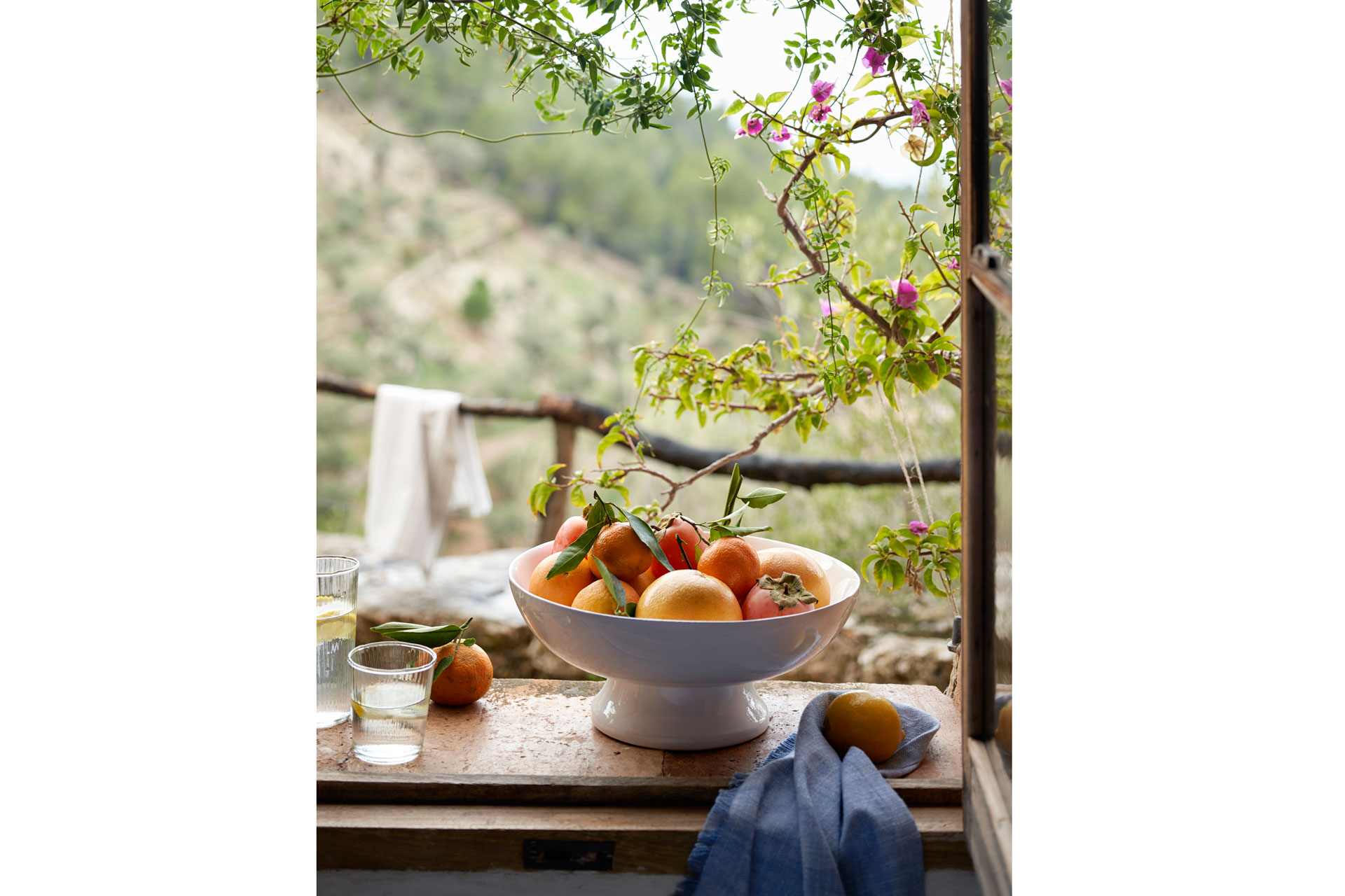a fruit bowl of orangs sits by a window overlooking a Mediterranean landscape