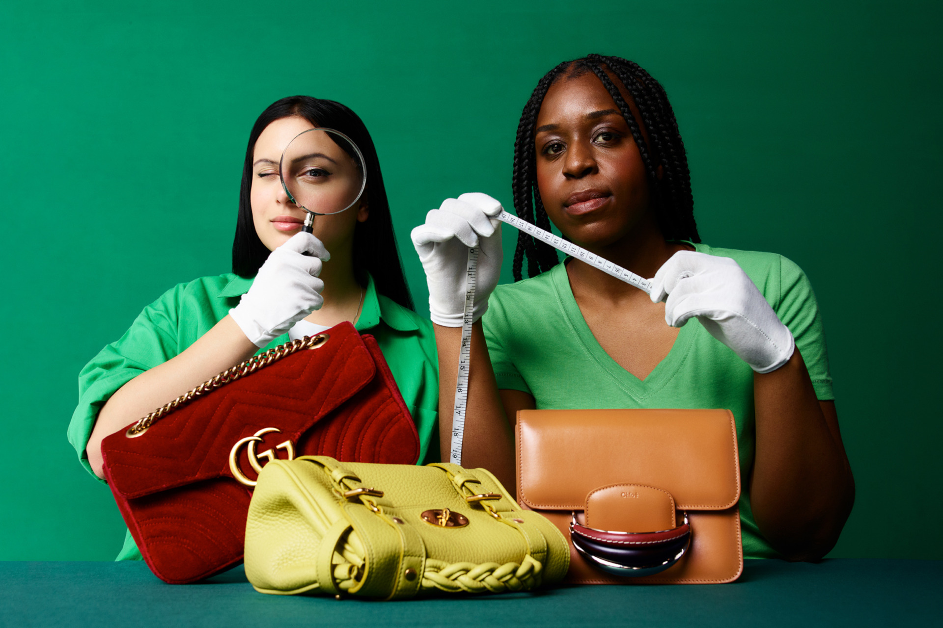 Two women holding measuring tape and a magnifying glass to inspect handbags on the table in front of them
