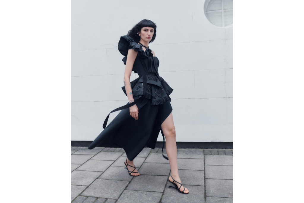 Model in black one-shoulder dress walking past with her head turned towards the camera