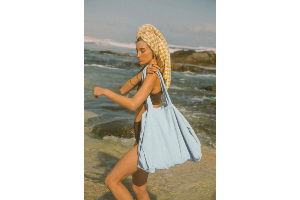 Model on the beach with hair wrapped in a towel pulling a blue bag over her shoulder