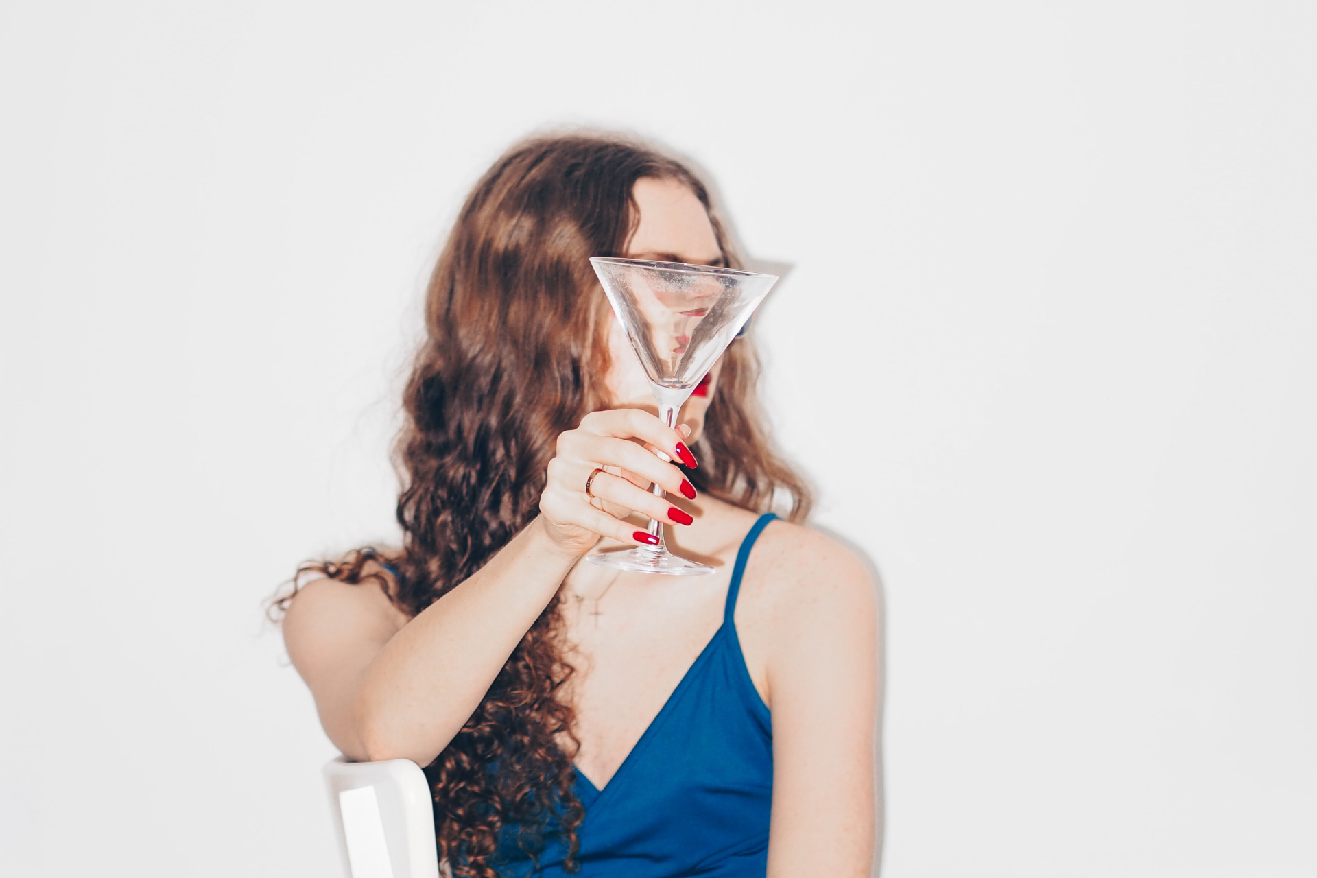 Redhaired woman in blue dress holding martini glass in front of her face, as she sits on a white chair in front of a white background