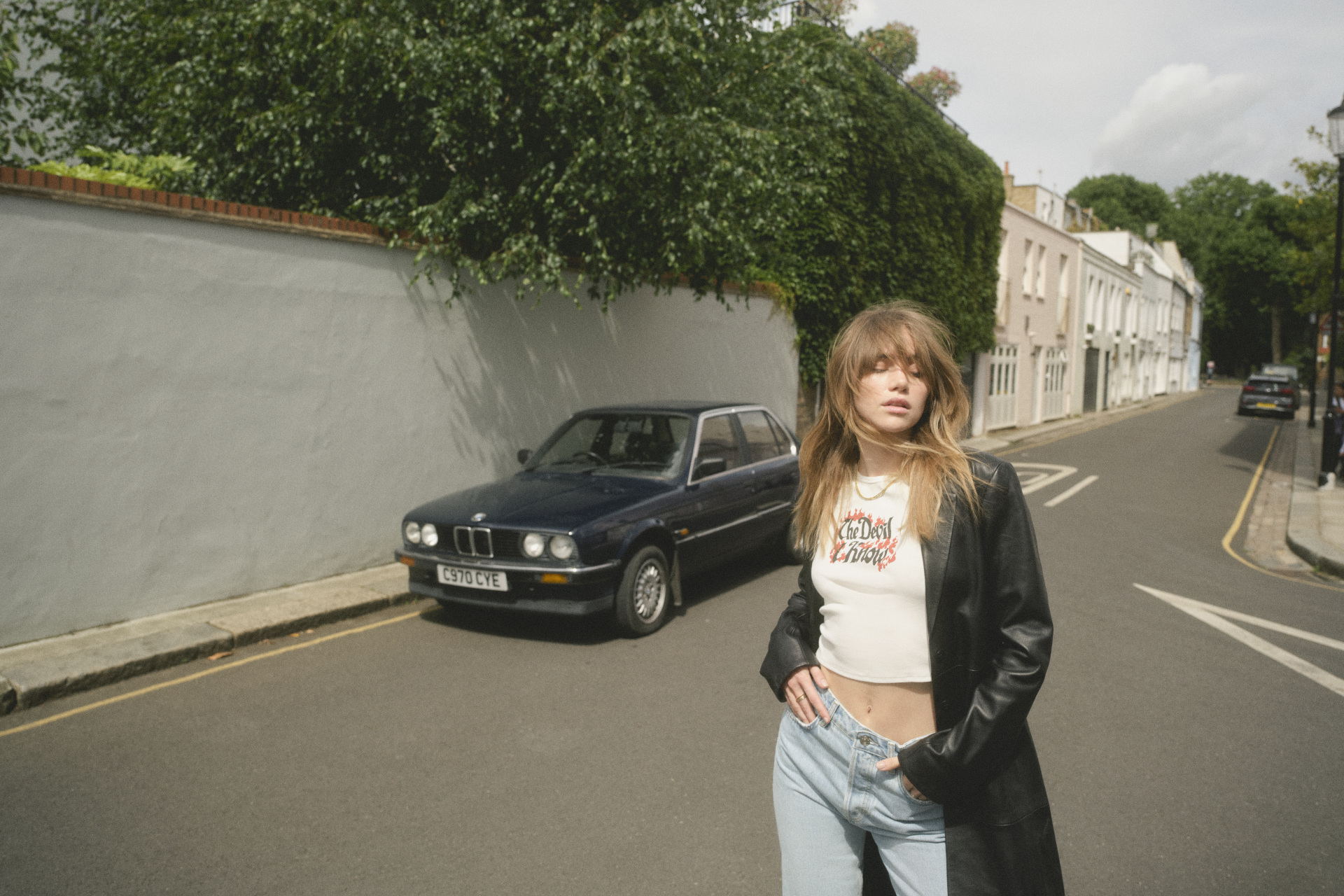 Suki Waterhouse wearing a white graphic tee, long black jacket and jeans, stood in the middle of a London neighbourhood