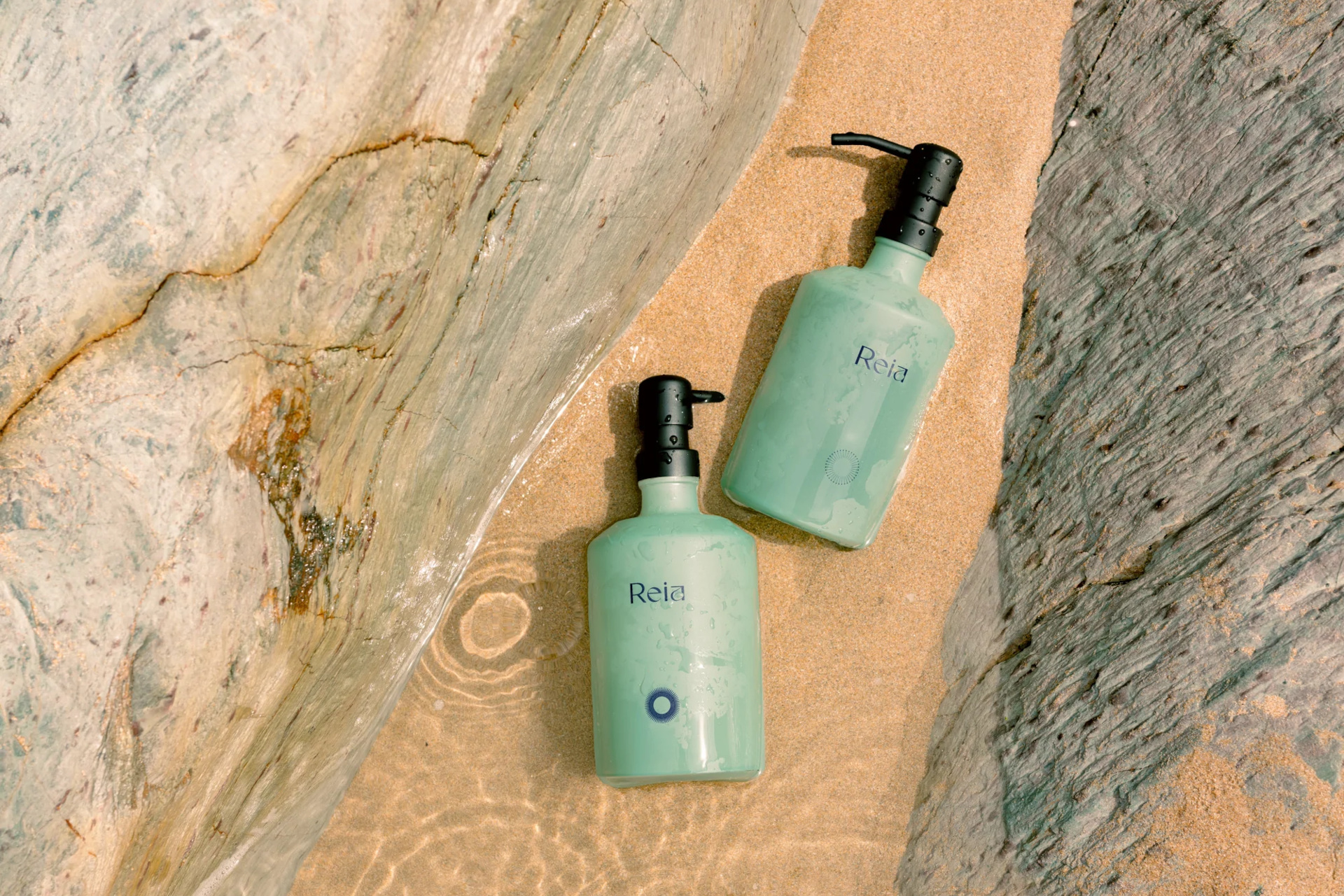 Two green bottles sat in the sand between two rocks, with water flowing through the space