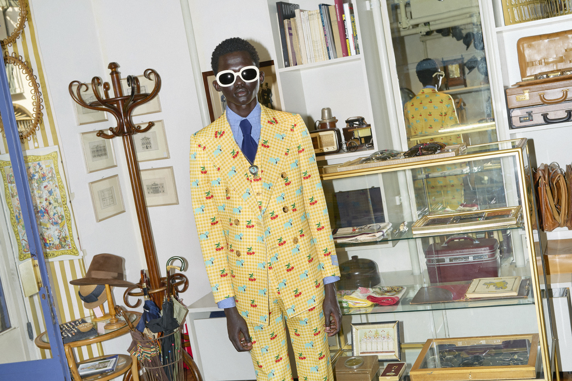 Black male model stood in antiques and curios shop, wearing a yellow printed suit and large black sunglasses with white frames
