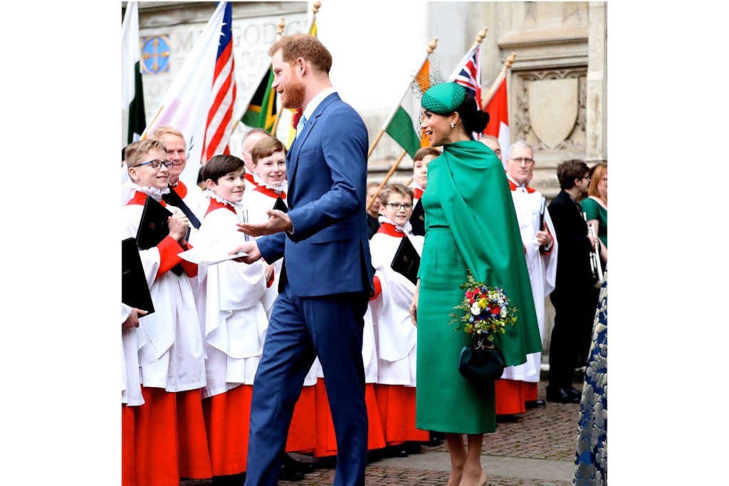 Meghan Markle and Prince Harry walking past choir boys, with flags in the background. Meghan is wearing a green midi dress with a matching cape and hat, Harry is wearing a blue suit