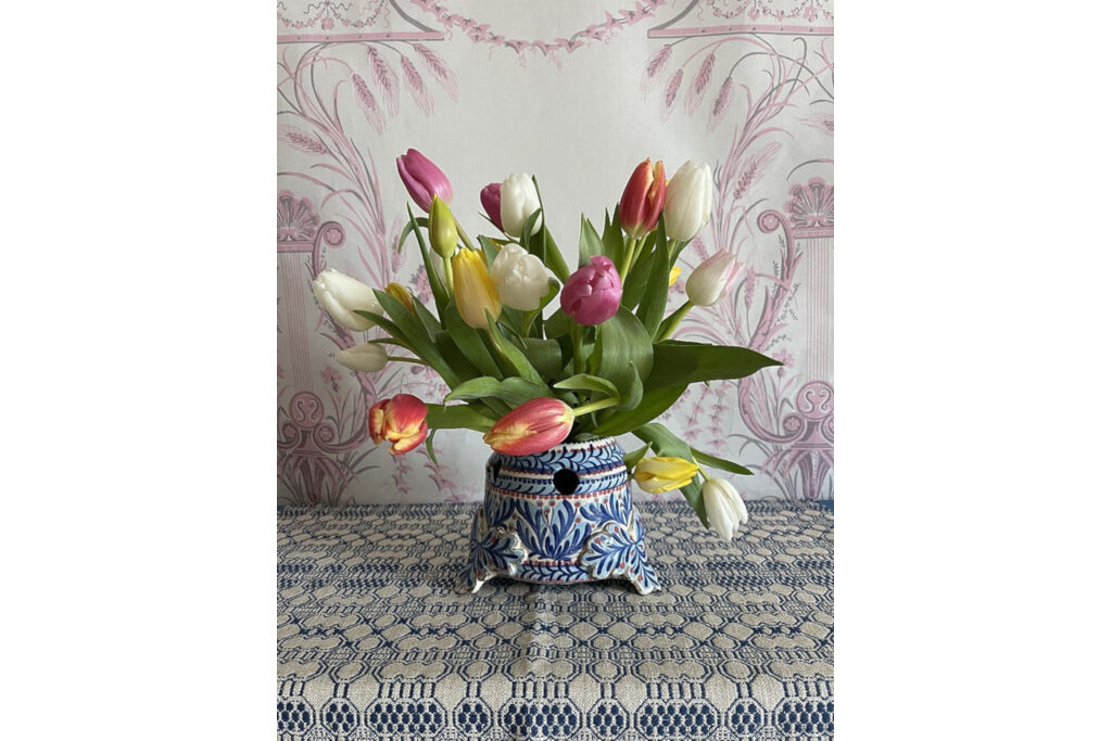 Tulipiere vase with tulips sat on patterned tablecloth against patterned wall