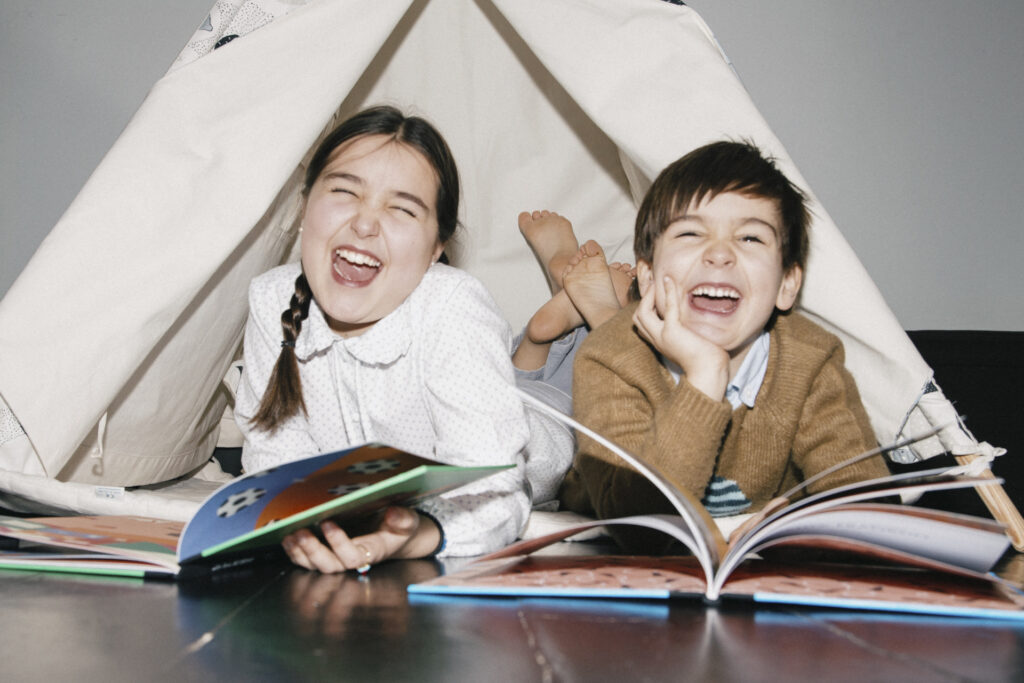 A girl and a boy in a cloth tent set up indoors, laying down on their fronts with books open in front of them as they smile at the camera