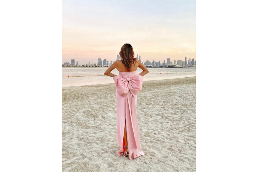 Woman on the beach facing away from the camera, wearing a pink evening dress with a large bow on the back