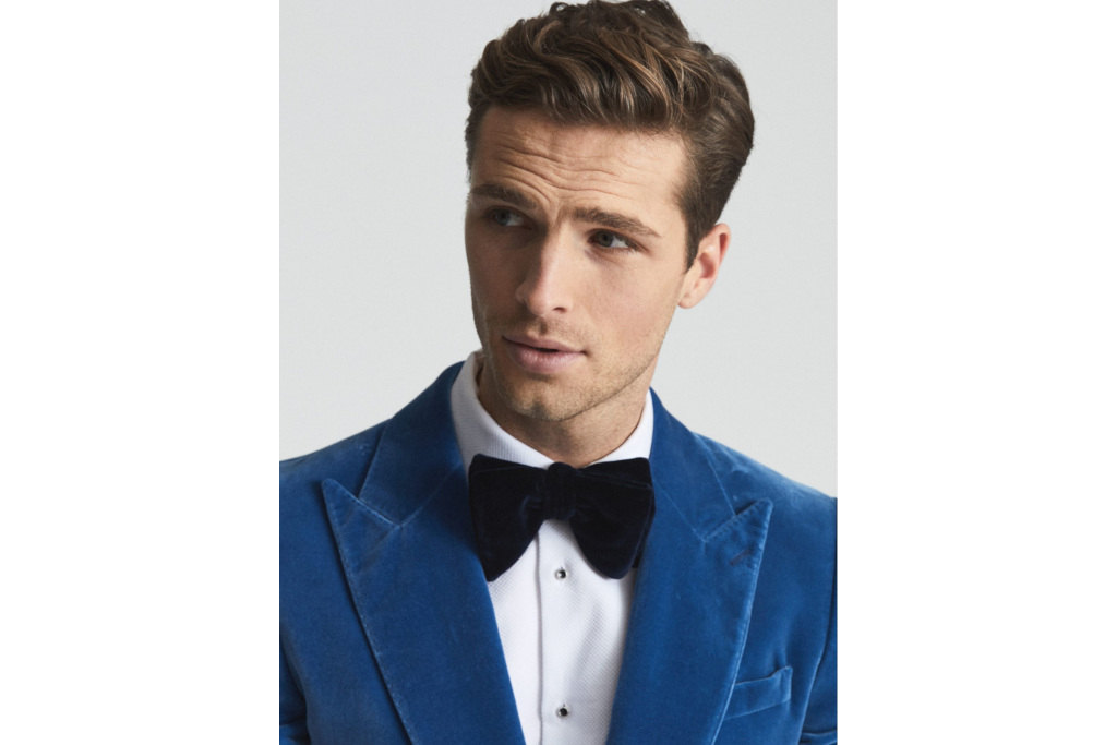 Close up of a man in a blue velvet blazer, white shirt and black velvet bow tie