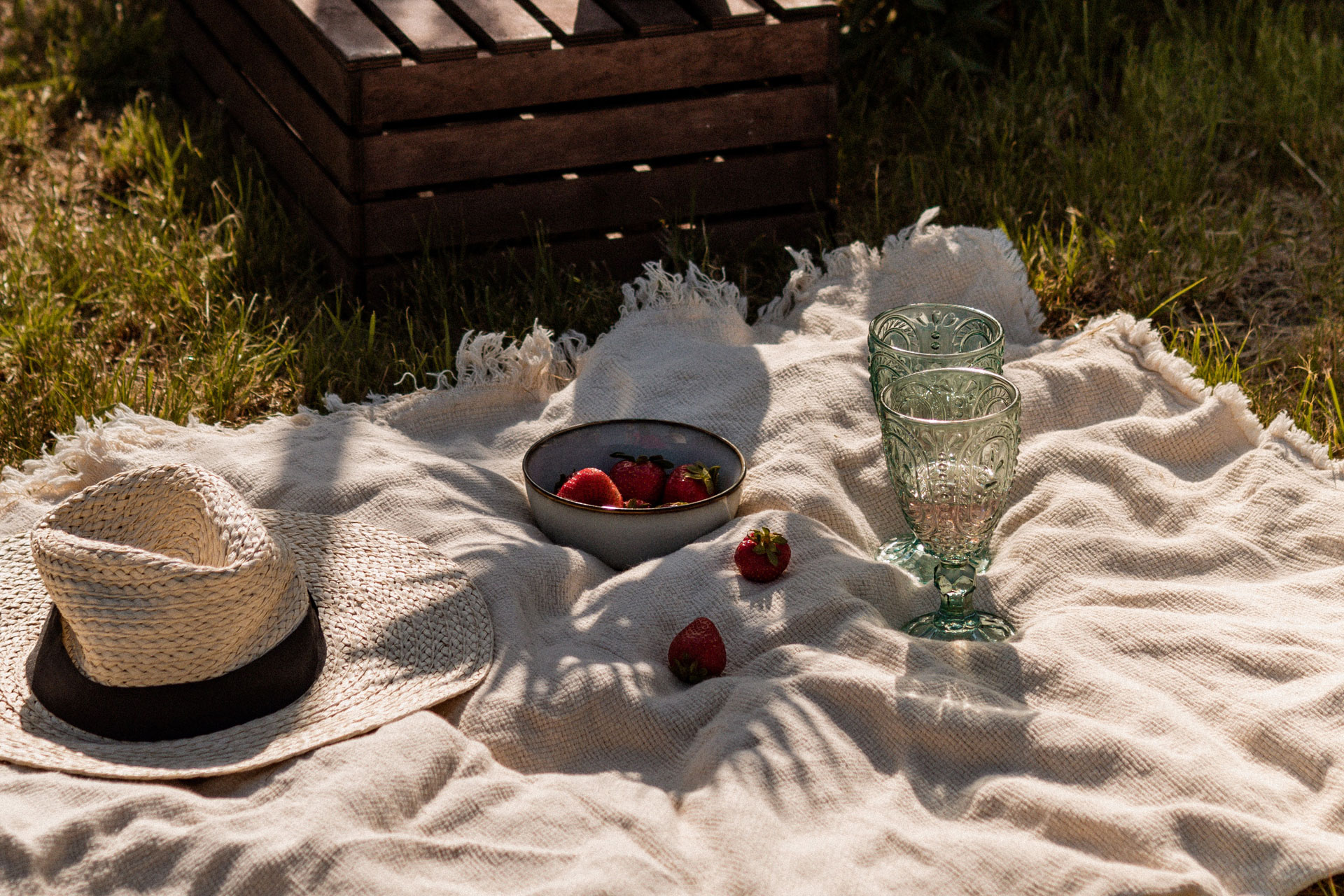 a picnic blanket with two glasses, a sun hat and a bowl of strawberries