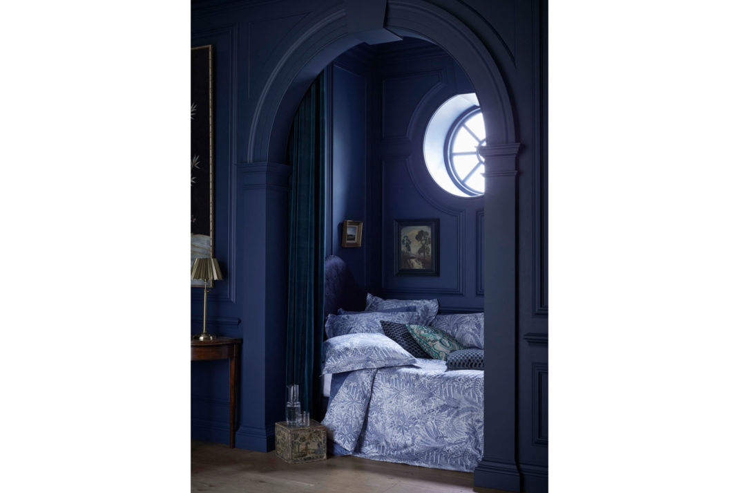 a dark blue bedroom with a l'oiel de boeuf window letting light in, with liberty bedding and pillows featuring