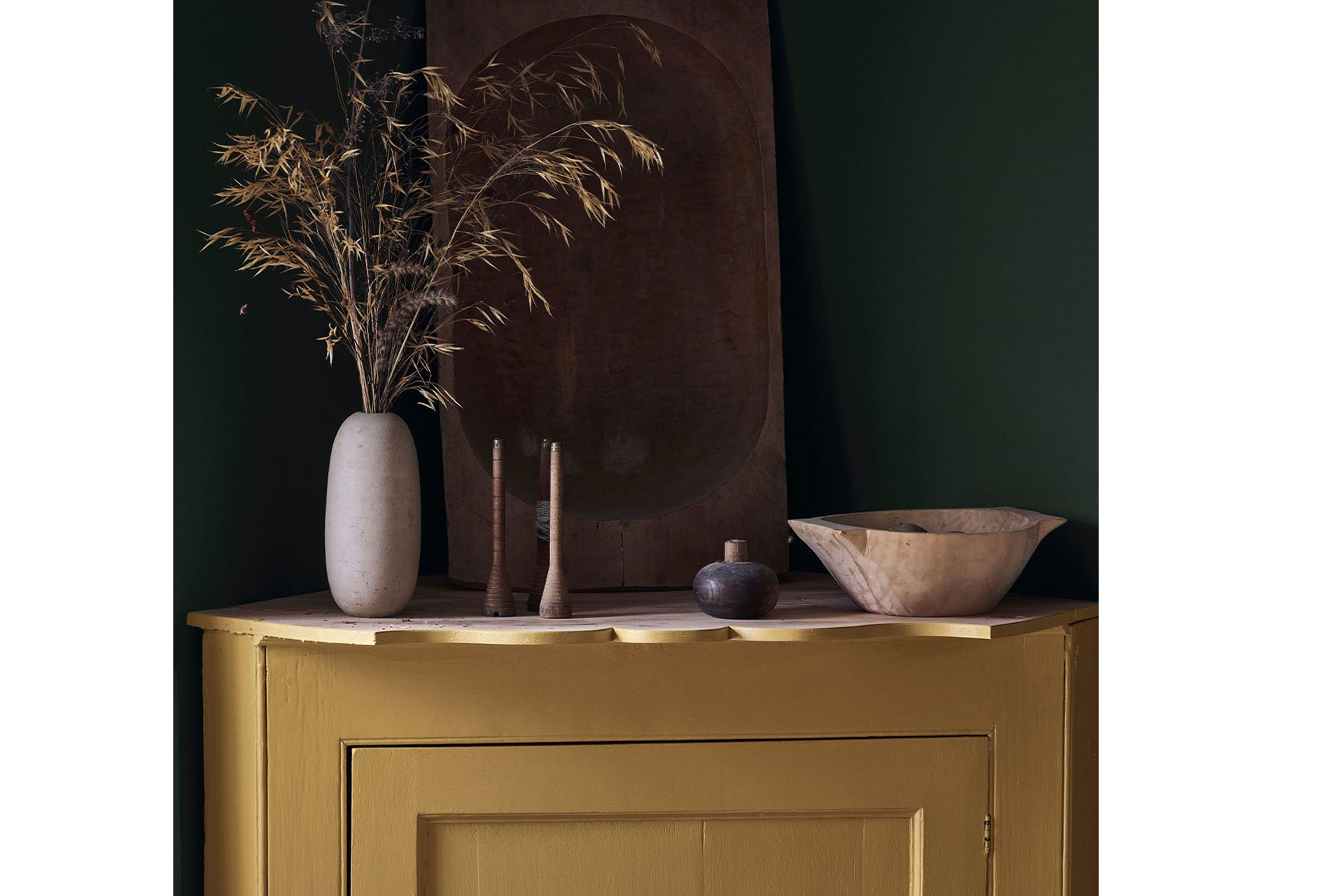 a yellow dresser with natural dried plants stands in front of a dark green wall