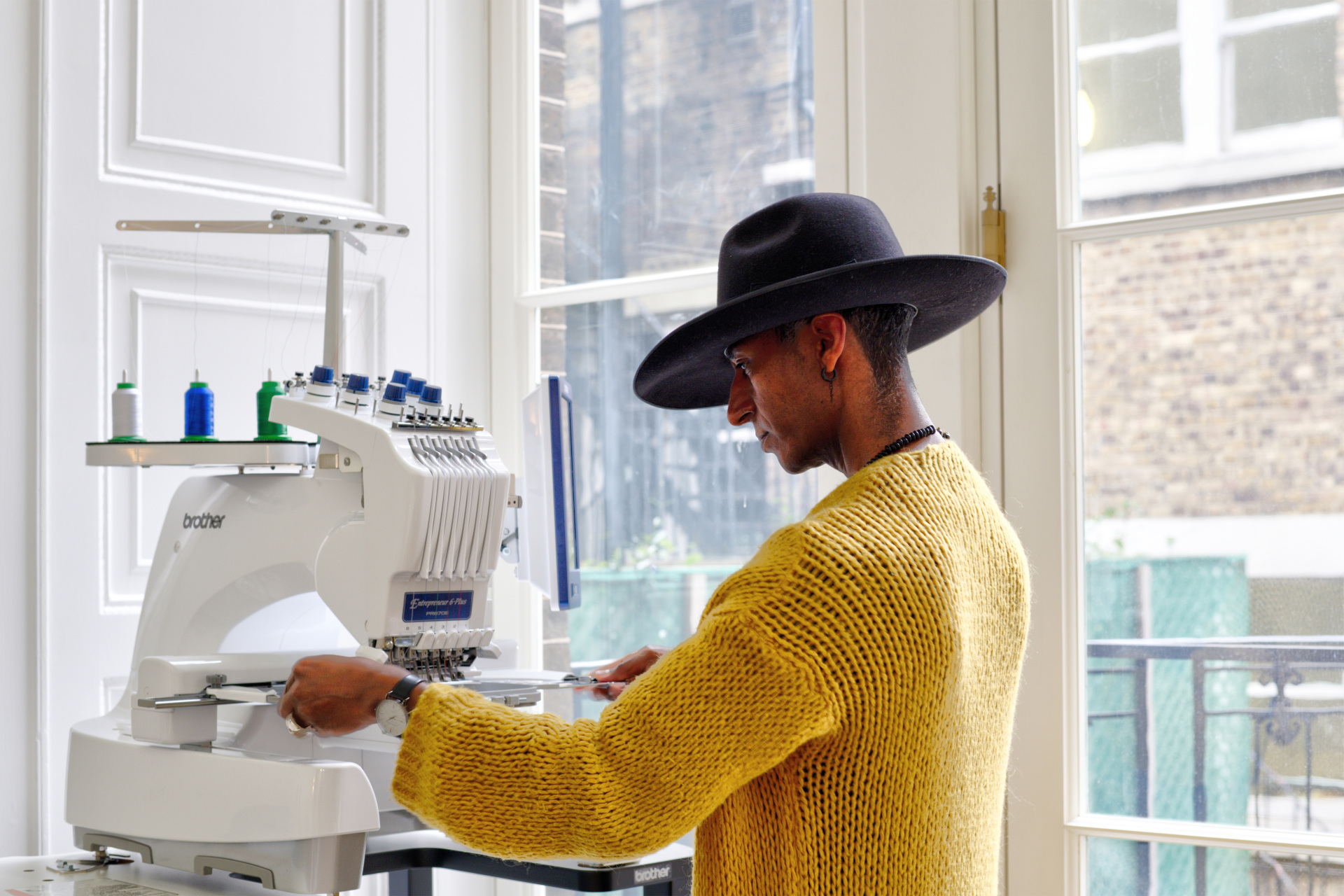 Person in a yellow jumper and wide brim hat pushing fabric through a sewing machine
