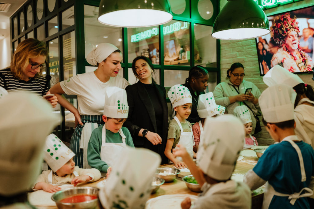 Children in chef hats making pizza as their parents look over them