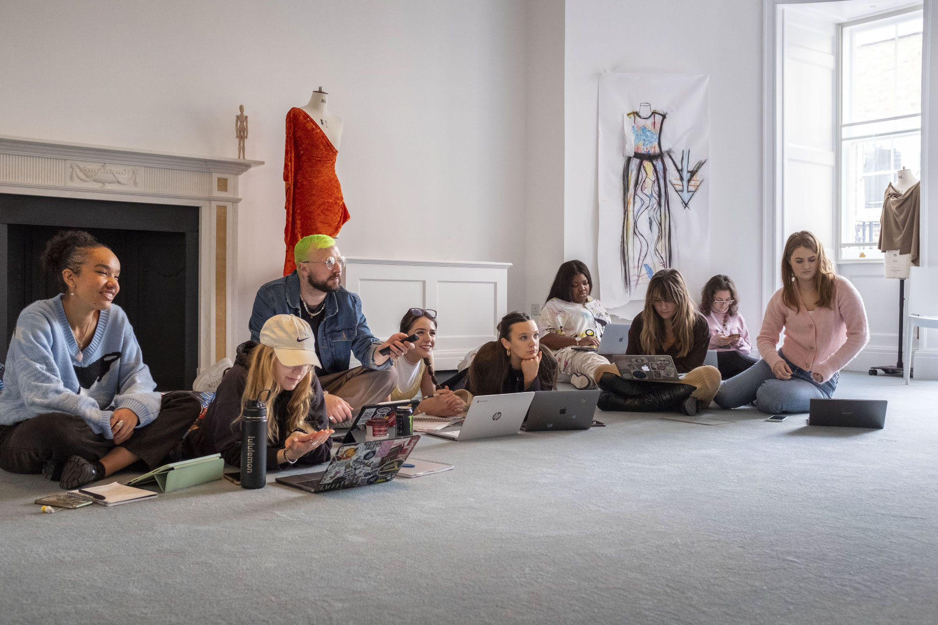 A group of students sat on the floor together with laptops, with mannequins in the background