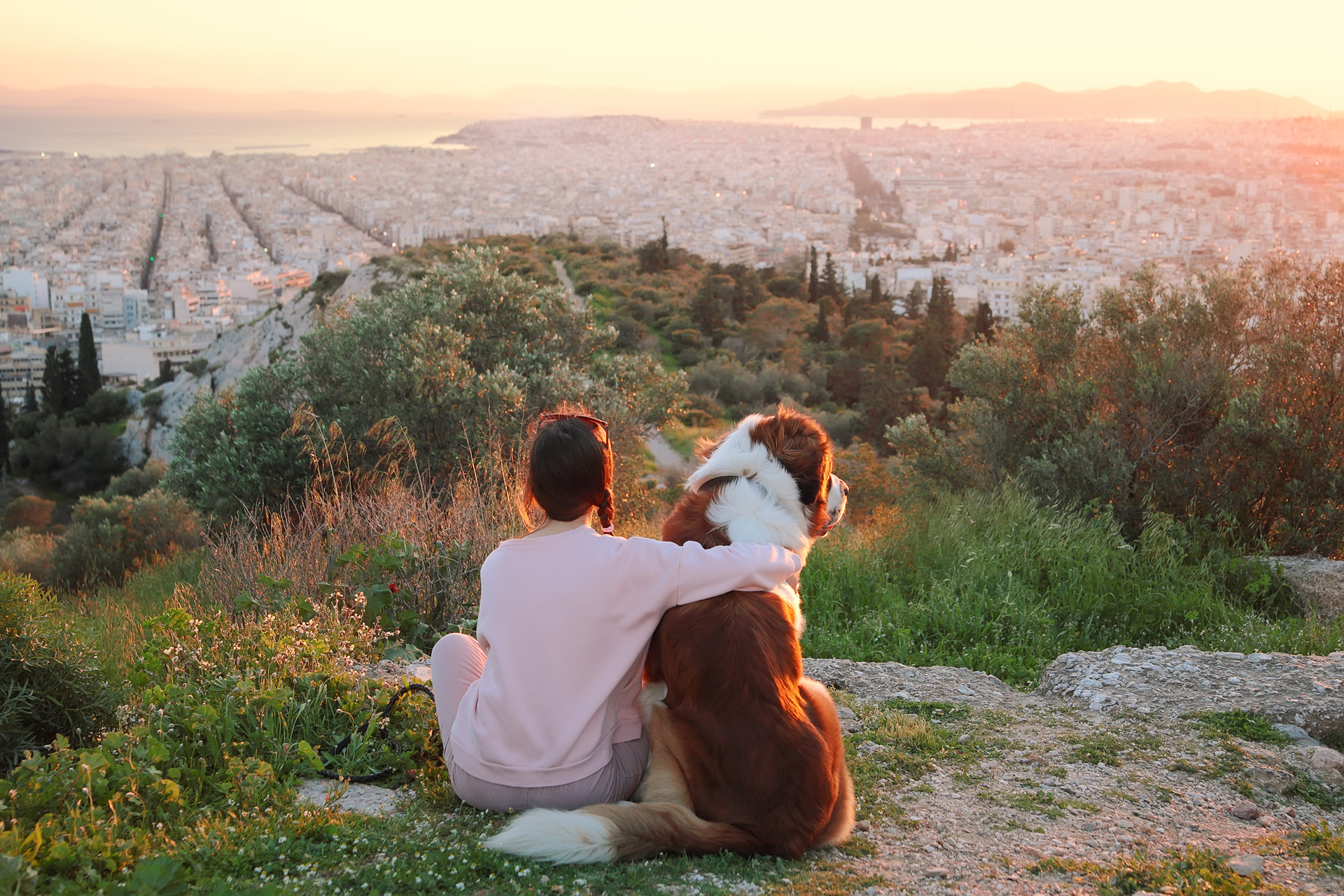 Young woman hugs her dog as they sit in a field. Athens, Greece