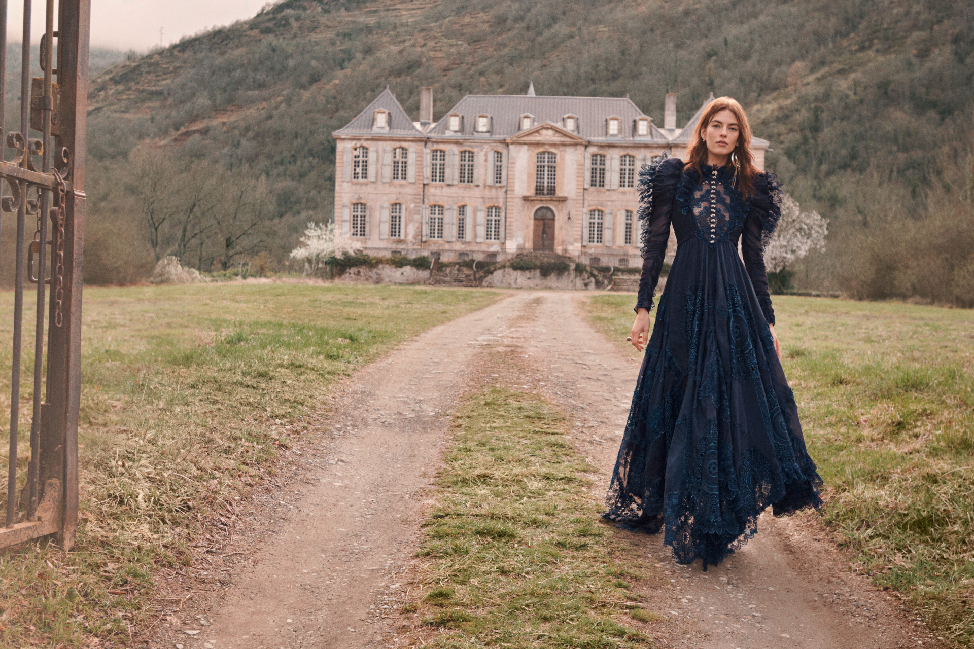 Model in long black dress walking towards camera, with an old country estate behind her
