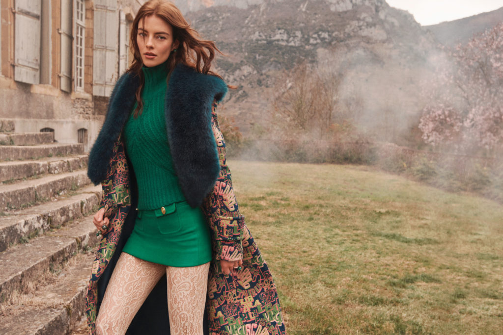 Model in a garden wearing a green dress, white lace tights and large floral jacket with fluffy green lapels