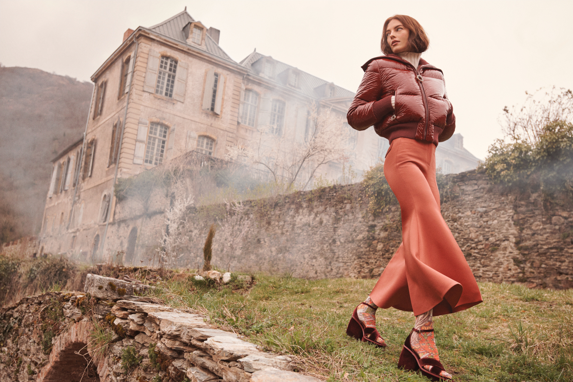 Model walking beside large country estate wearing a brown puffer jacket and coral-coloured trousers