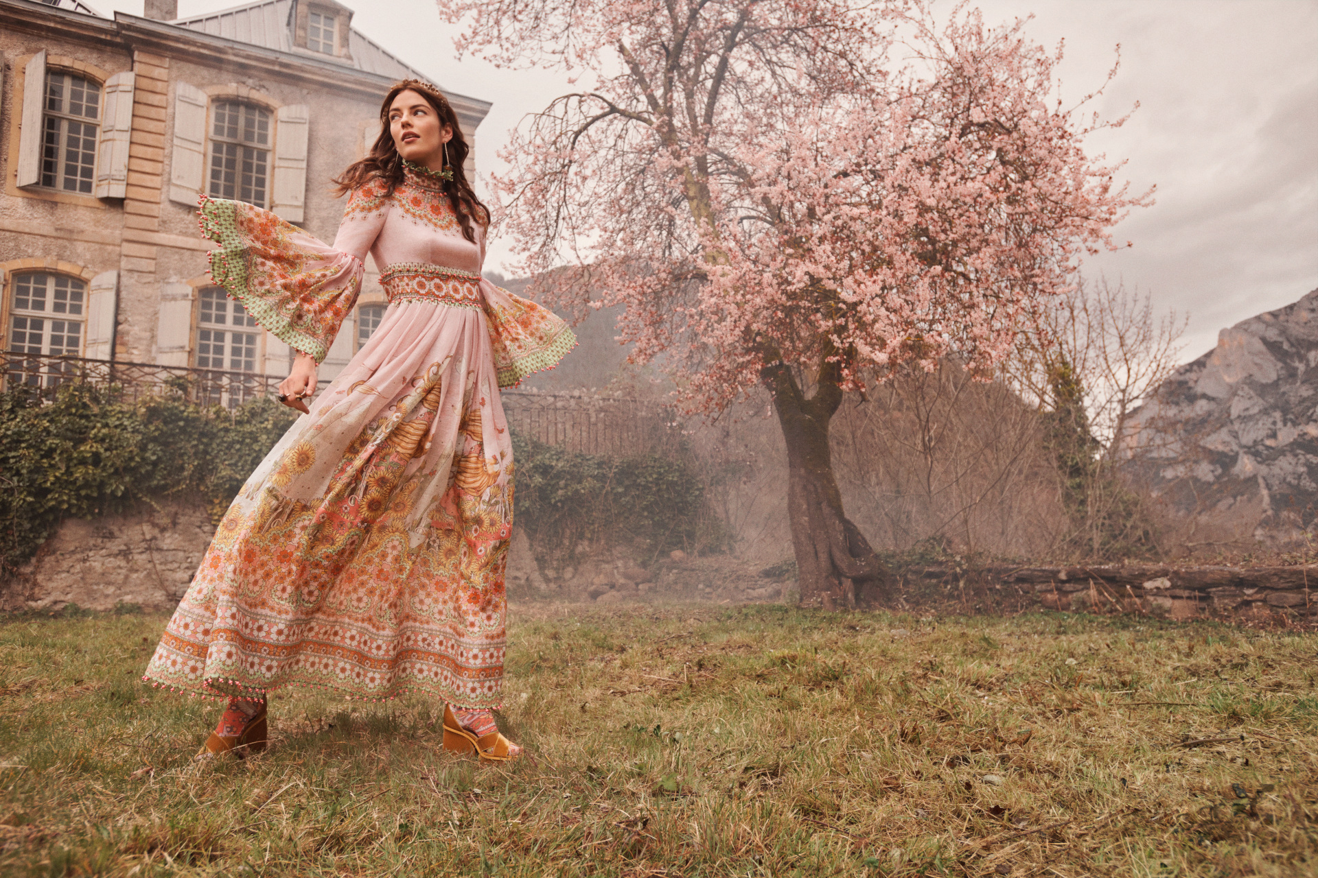 Model in garden wearing a long pink floral dress with a cherry blossom tree in the background